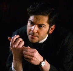 Daniel Maslany as Llewellyn Watts in Murdoch Mysteries, a white man with dark curly hair and brown eyes, leans forward with furrowed eyebrows while fidgeting worriedly with his hands.