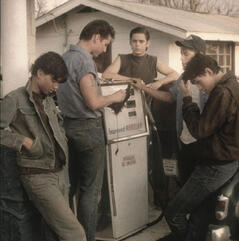 A candid photo of The Outsiders movie adaption cast gathered around a gas pump