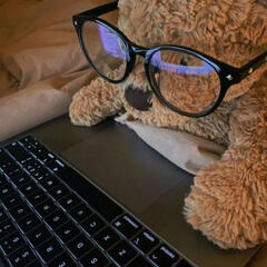 A brown teddy bear lays on a bed, propped up next to an open laptop as though using it. a pair of eyeglasses are situated on its face to further the illusion.