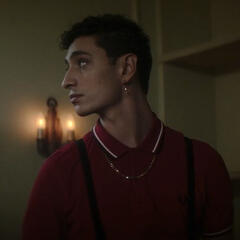 Jayden Revri as Charles Rowland in Dead Boy Detectives, an Indian-Jamaican man with short dark curls and dark eyes, looks off to the left with a blank facial expression, as though responding to his name being called. He wears a red polo shirt and gold jewe